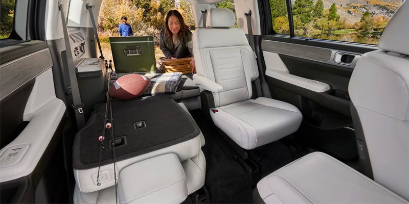 A woman sits in the back of a large SUV with a basket, alongside camping gear and outdoor equipment.