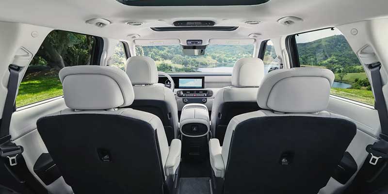 A spacious car interior shows rows of gray seats and a view of a green, hilly landscape through the windshield.